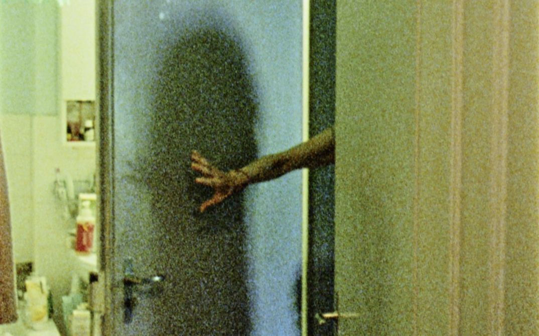 Filmstill aus "Spuren von Bewegung vor dem Eis" von René Frölke. Zu sehen ist ein Badezimmer. Die Badezimmertür wird von einem Arm offen gehalten.