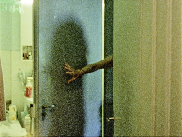 Film still from "Spuren von Bewegung vor dem Eis" by René Frölke. It shows a bathroom. The bathroom door is held open by an arm. 