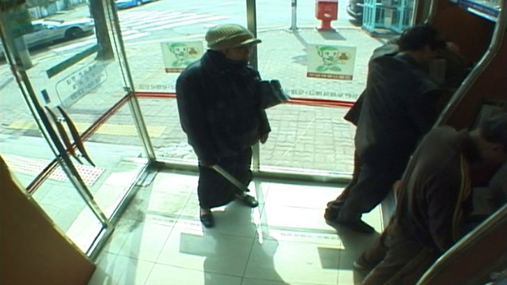 Film still from INFLUENZA: You see the recording from a surveillance camera, people at ATMs.