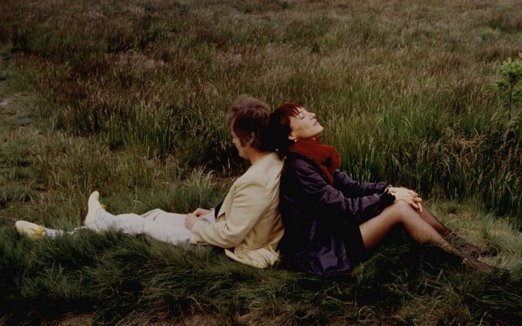 Filmstill aus DER BEGINN ALLER SCHRECKEN IST LIEBE. Rücken an Rücken sitzen ein Mann und eien Frau im Gras.