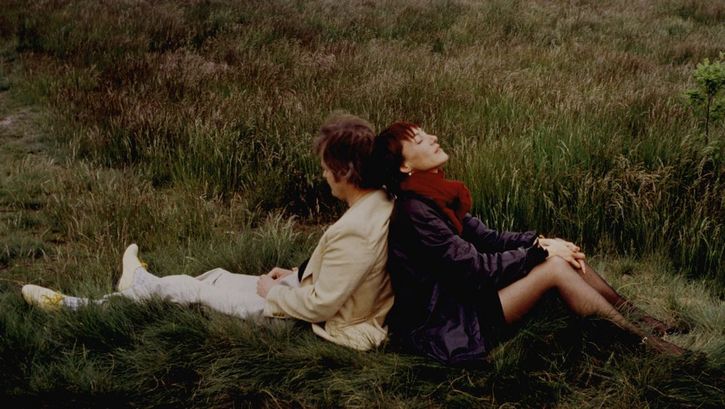 Filmstill aus DER BEGINN ALLER SCHRECKEN IST LIEBE. Rücken an Rücken sitzen ein Mann und eien Frau im Gras.