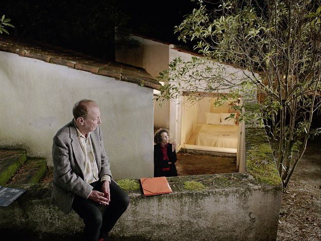 Filmstill aus „O trio em mi bemol (The Kegelstatt Trio)“ von Rita Azevedo Gomes. Ein Mann sitzt auf einer Gartenmauer. Im Hintergrund steht eine Frau am Fuß einer Treppe und raucht. 