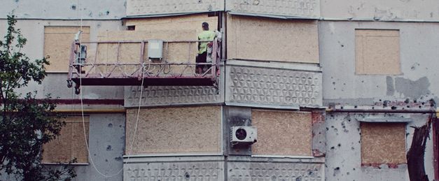 It shows the wall of a building. A man on a lifting platform covers the windows with wooden boards.