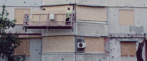 Zu sehen ist die Wand eines Gebäudes. Ein Mann auf einer Hebebühne bedeckt die Fenster mit Holzbrettern.