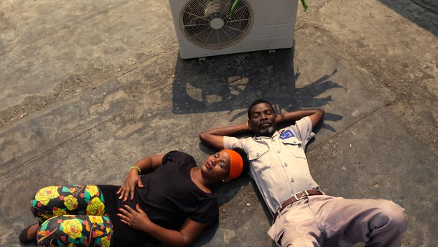 Film still from AIR CONDITIONER: A man and a woman lie next to each other on the floor, next to them is a fan.