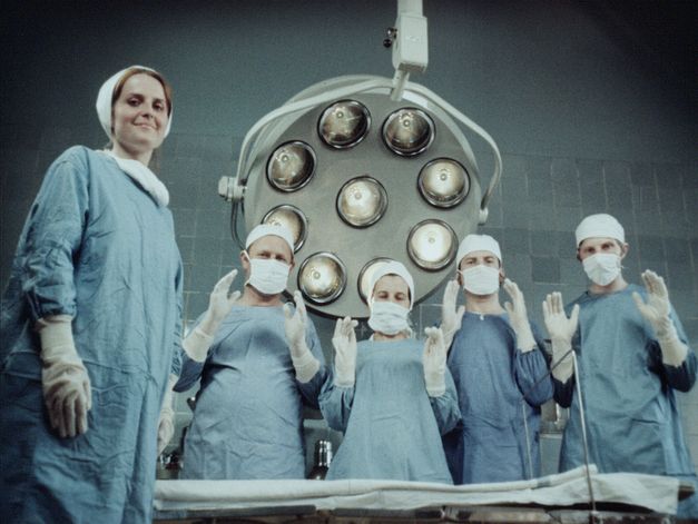 Film still from Zuza Banasińska’s film “Grandmamauntsistercat”. Five nurses wear scrubs and look down at the camera. One of them smiles, whilst the other four, also wearing facemasks, hold both their hands up in front of their chests. A large machine can be seen in the background.