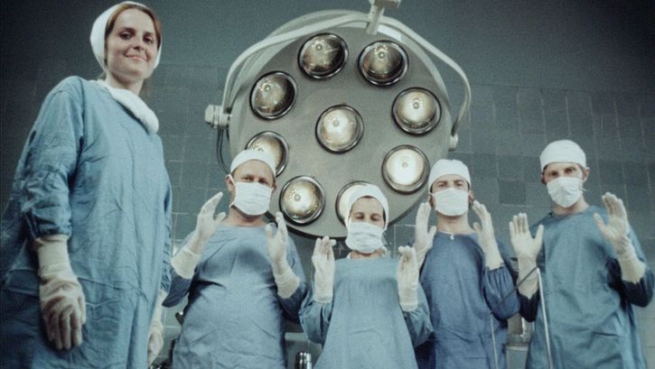 Film still from Zuza Banasińska’s film “Grandmamauntsistercat”. Five nurses wear scrubs and look down at the camera. One of them smiles, whilst the other four, also wearing facemasks, hold both their hands up in front of their chests. A large machine can be seen in the background.
