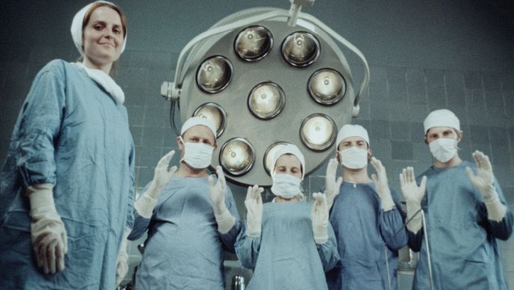 Film still from Zuza Banasińska’s film “Grandmamauntsistercat”. Five nurses wear scrubs and look down at the camera. One of them smiles, whilst the other four, also wearing facemasks, hold both their hands up in front of their chests. A large machine can be seen in the background.
