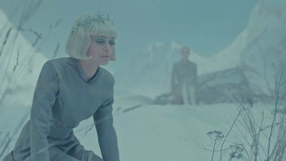 Filmstill aus LA TOUR DE GLACE: In einer Schneelandschaft blickt eine märchenhaft gekleidete Frau vor sich hin. Im Hintergrund sieht man verschwommen Berge und eine weitere Person.