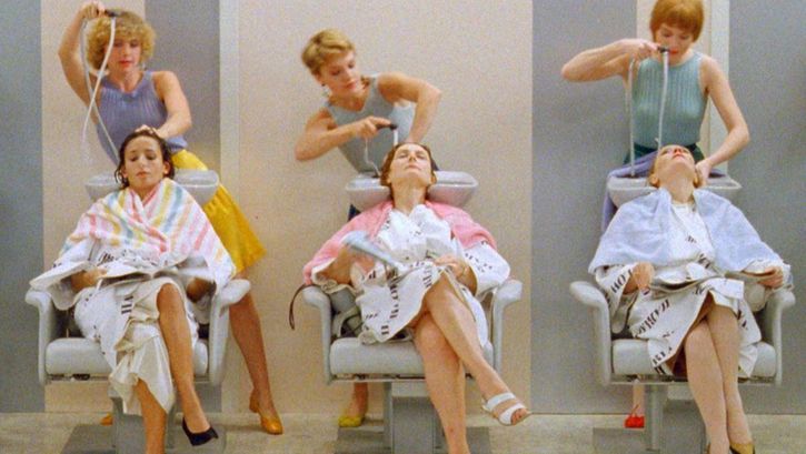 Filmstill aus GOLDEN EIGHTIES: Drei Frauen sitzen in einer Reihe in Friseurstühlen. Hinter ihnen steht je eine Friseurin und wäscht ihnen die Haare. Alle sind sommerlich gekleidet.