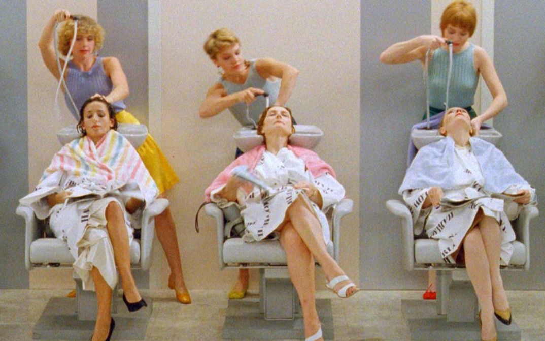 Filmstill aus GOLDEN EIGHTIES: Drei Frauen sitzen in einer Reihe in Friseurstühlen. Hinter ihnen steht je eine Friseurin und wäscht ihnen die Haare. Alle sind sommerlich gekleidet.