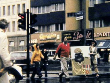 Filmstill aus „Aufenthaltserlaubnis" von Antonio Skármeta. Altes Bild von Menschen, die auf der Straße laufen, und eine Karikatur von Idi Amin tragen.
