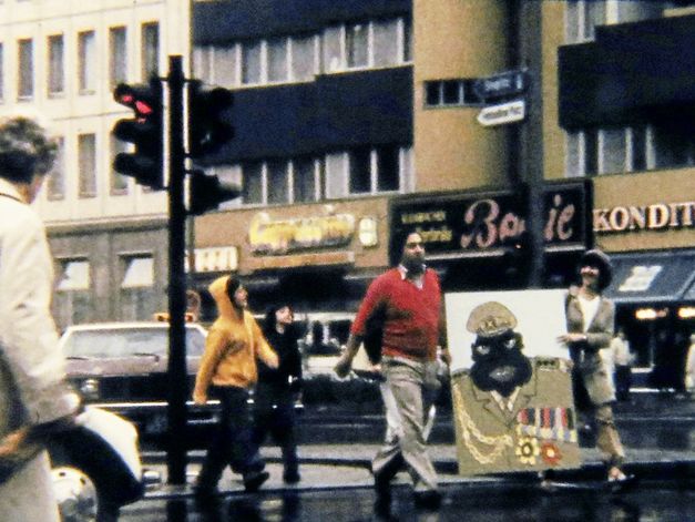 Filmstill aus „Aufenthaltserlaubnis" von Antonio Skármeta. Altes Bild von Menschen, die auf der Straße laufen, und eine Karikatur von Idi Amin tragen.