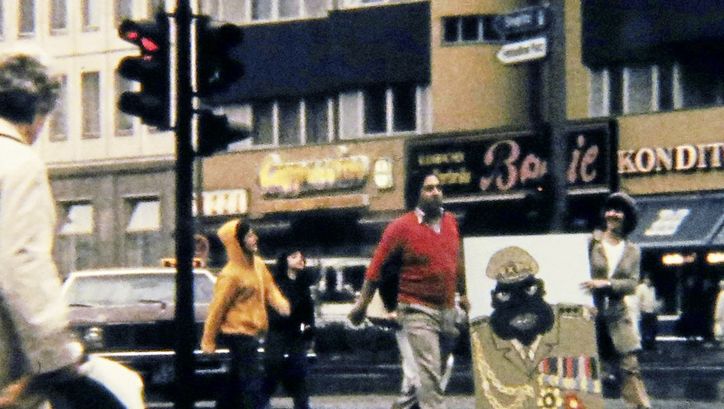 Filmstill aus „Aufenthaltserlaubnis" von Antonio Skármeta. Altes Bild von Menschen, die auf der Straße laufen, und eine Karikatur von Idi Amin tragen.
