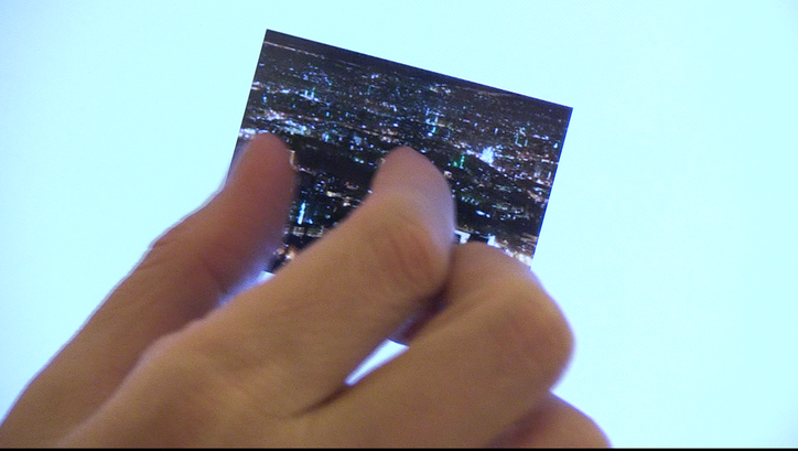 Film still from IN FIELDS OF WORD: CONVERSATIONS WITH SAMAR YAZBEK: A hand holds a picture in front of the sky.