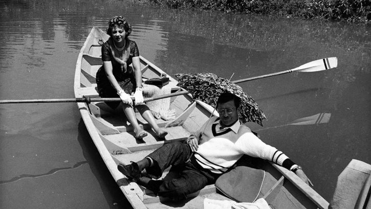 Film still from LA VISITA: A man and a woman are sitting in a rowing boat. She is rowing, he is protecting himself from the sun with an umbrella.