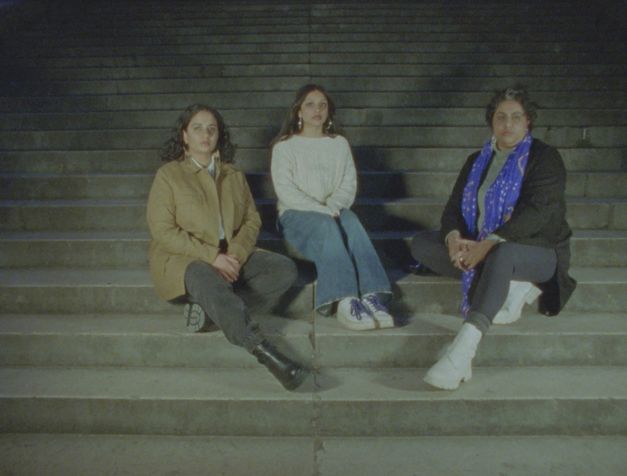 Film still from Cana Bilir-Meier’s “Zwischenwelt (In-between World)”. Three people sit on a staircase and look directly at us. 