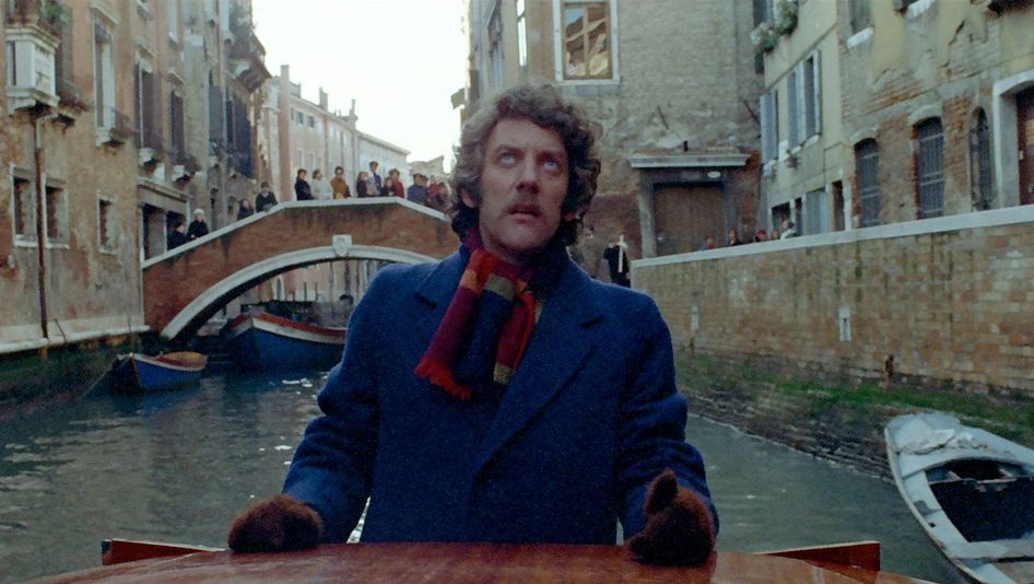 Film still from DON'T LOOK NOW: A man is standing in a gondola in a canal in Venice, looking up. In the background, a bridge with people on it can be seen.