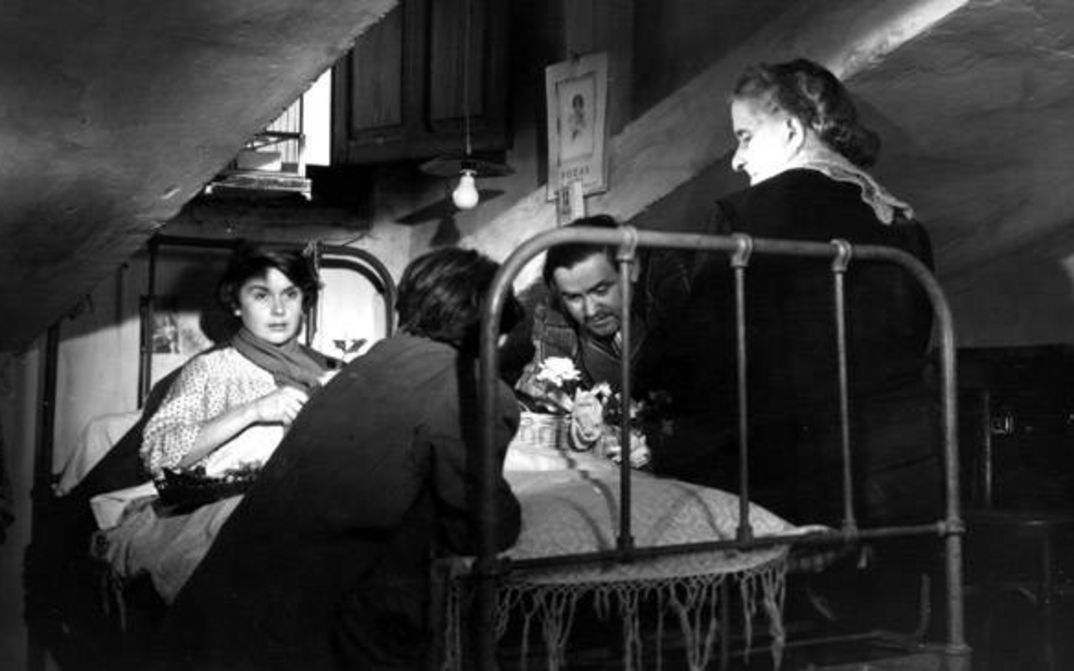 Film still from SEGUNDO LOPEZ, AVENTURERO URBANO: A sick young woman is lying in bed in an attic. Three people are standing around the bed.