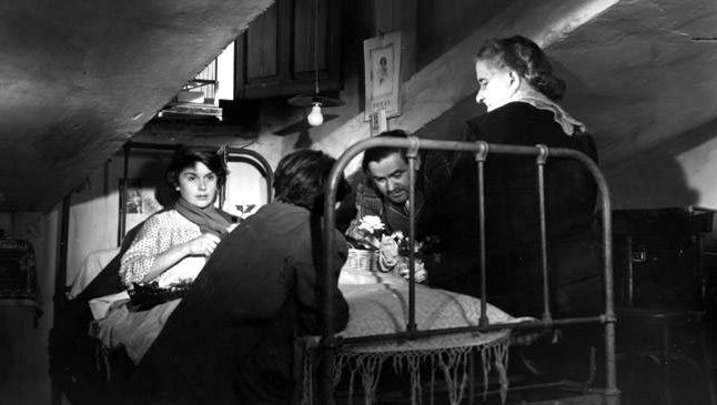 Film still from SEGUNDO LOPEZ, AVENTURERO URBANO: A sick young woman is lying in bed in an attic. Three people are standing around the bed.