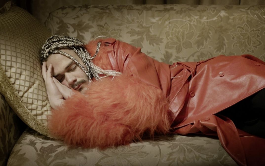 Filmstill aus „Llamadas desde Moscú“ von Luís Alejandro Yero. Ein Mann in einem roten Mantel schläft auf einem Sofa.