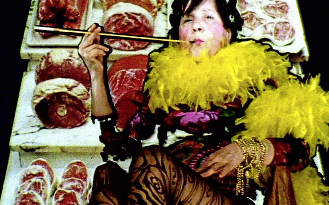 Filmstill aus "Maria Lassnig Kantate" von Maria Lassnig, Hubert Sielecki. Zu sehen ist eine auffällig gekleidete Person mit einer Federboa und einer Zigarettenspitze. Der Hintergrund besteht aus Thekenfleisch.