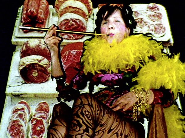 Filmstill aus "Maria Lassnig Kantate" von Maria Lassnig, Hubert Sielecki. Zu sehen ist eine auffällig gekleidete Person mit einer Federboa und einer Zigarettenspitze. Der Hintergrund besteht aus Thekenfleisch.