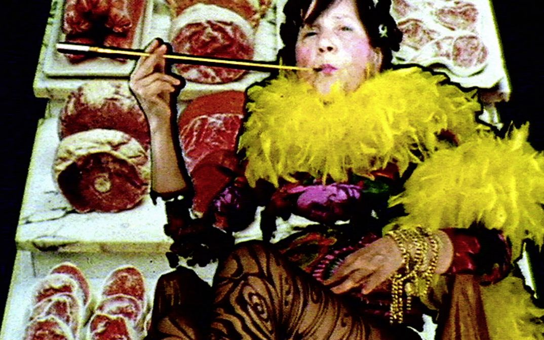 Filmstill aus "Maria Lassnig Kantate" von Maria Lassnig, Hubert Sielecki. Zu sehen ist eine auffällig gekleidete Person mit einer Federboa und einer Zigarettenspitze. Der Hintergrund besteht aus Thekenfleisch.