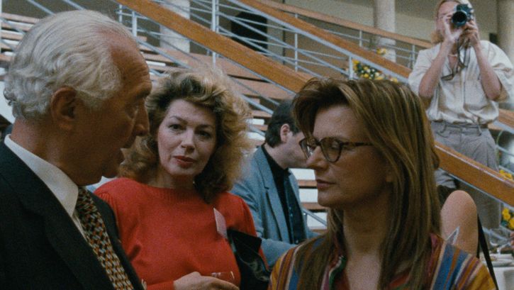 Filmstill aus DIE DEUTSCHEN UND IHRE MÄNNER. BERICHT AUS BONN: Drei Personen unterhalten sich miteinander und trinken Wein. Im Hintergrund eine Treppe und eine Person, die von oben fotografiert. 