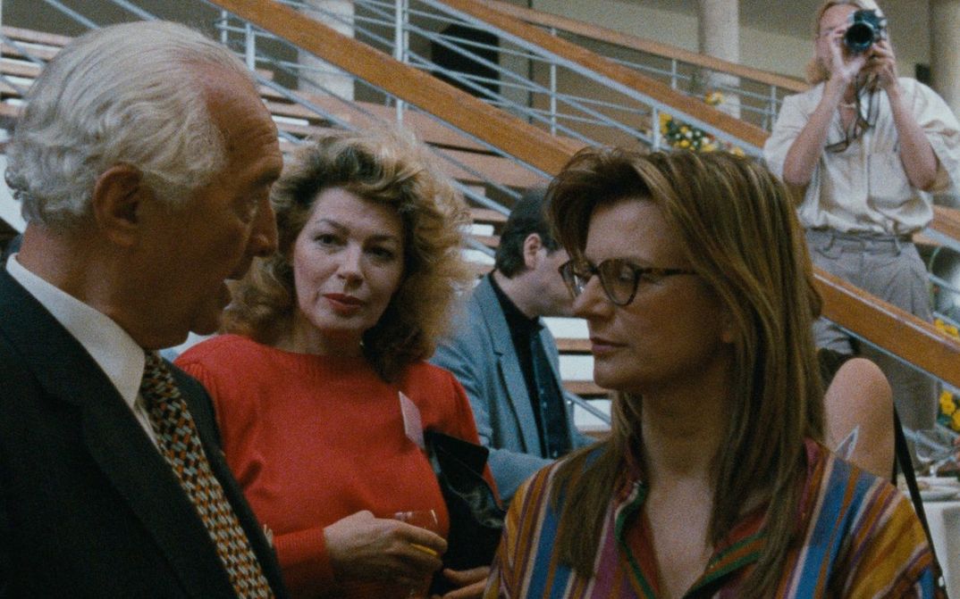 Film still from DIE DEUTSCHEN UND IHRE MÄNNER. REPORT FROM BONN: Three people are talking to each other and drinking wine. In the background a staircase and a person taking a photo from above. 