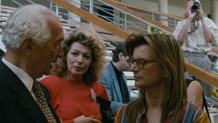 Filmstill aus DIE DEUTSCHEN UND IHRE MÄNNER. BERICHT AUS BONN: Drei Personen unterhalten sich miteinander und trinken Wein. Im Hintergrund eine Treppe und eine Person, die von oben fotografiert. 