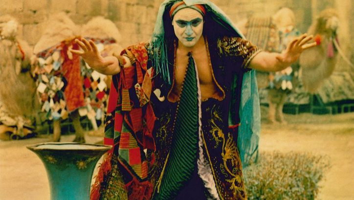 Film still from DIE LEGENDE DER FESTUNG SURAMI: A man in a colorful disguise with a painted face raises his hands imploringly.