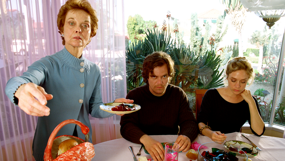 Filmstill aus MY SON, MY SON, WHAT HAVE YE DONE: Drei Menschen am Esstisch. Eine ältere Frau hält stehend einen Teller in der Hand und zeigt mit der anderen Hand auf etwas außerhalb des Bildausschnittes. 