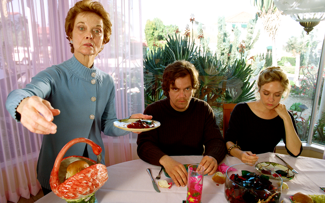 Film still from MY SON, MY SON, WHAT HAVE YE DONE: Three people at the dining table. An older woman is standing, holding a plate in her hand and pointing with her other hand to something outside the frame.