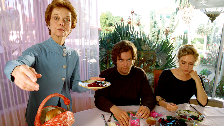 Film still from MY SON, MY SON, WHAT HAVE YE DONE: Three people at the dining table. An older woman is standing, holding a plate in her hand and pointing with her other hand to something outside the frame. 