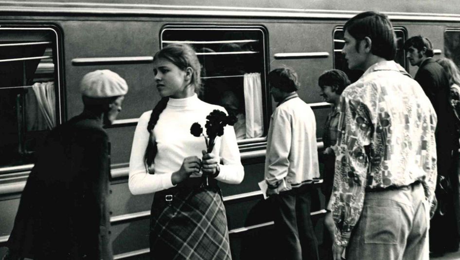 Filmstill aus DER SPECHT ZERBRICHT SICH NICHT DEN KOPF: Einige Menschen stehen an einem Bahnsteig, hinter sich ein stehender Zug. Ein junges Mädchen hält Blumen in der Hand und blickt sehnsuchtsvoll in die Ferne.