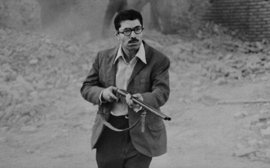Film still from POSTSCHI: A man wearing a shirt and jacket is standing on the street, pointing a rifle at something just outside the frame.