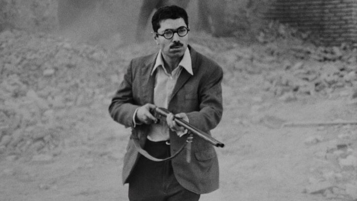 Film still from POSTSCHI: A man wearing a shirt and jacket is standing on the street, pointing a rifle at something just outside the frame.