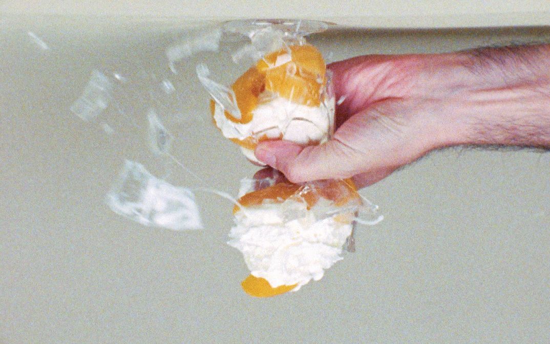Film still from PRINT ANALYSIS: A hand crushes a cream puff in a glass. The picture is upside down.