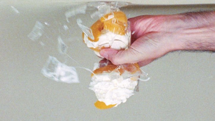 Film still from PRINT ANALYSIS: A hand crushes a cream puff in a glass. The picture is upside down.