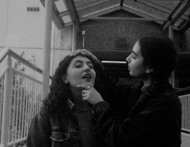 Still from the film "This Makes Me Want to Predict the Past" by Cana Bilir-Meier. A black and white image of two young women. One holds the head of the other, bends it slightly upwards and checks out something on her face.