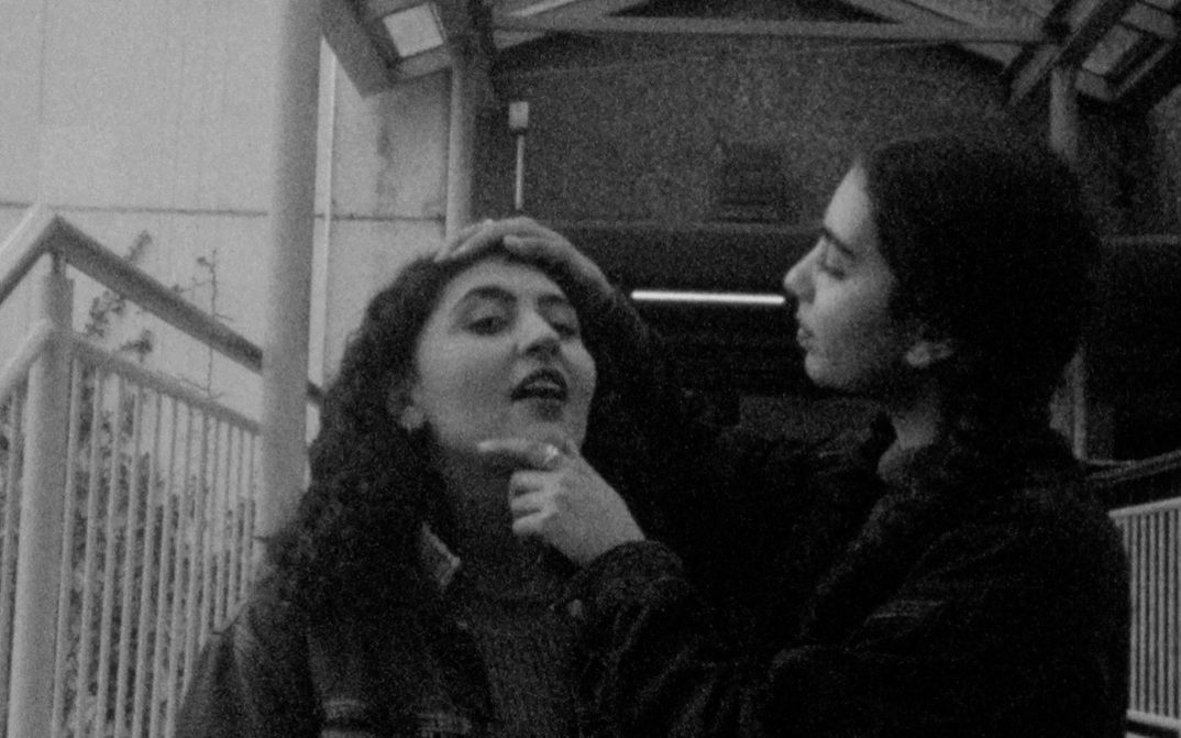 Filmstill aus „This Makes Me Want to Predict the Past“ von Cana Bilir-Meier. Ein Schwarz-Weiß-Bild von zwei jungen Frauen. Eine hält den Kopf der anderen hoch und schaut ihr ins Gesicht. 