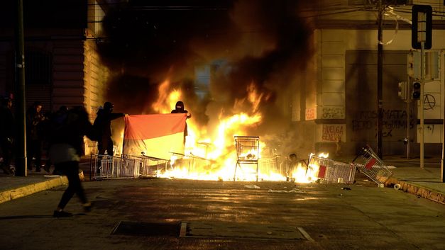 Filmstil aus "Oasis" von Tamara Uribe und Felipe Morgado. Zu sehen sind brennende Einkaufswagen auf der Straße und Menschen mit Masken. 