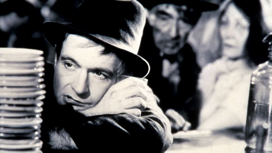 Film still from LA CHARETTE FANTÔME: A man wearing a hat leans on a bar counter, behind him other people can be seen out of focus.