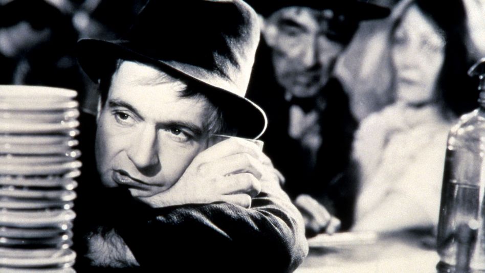Filmstill aus LA CHARETTE FANTÔME: Ein Mann mit einem Hut lehnt auf einem Bartresen, hinter ihm sind unscharf weitere Menschen zu sehen.