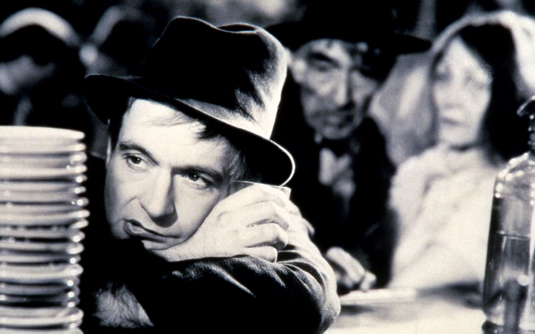 Film still from LA CHARETTE FANTÔME: A man wearing a hat leans on a bar counter, behind him other people can be seen out of focus.