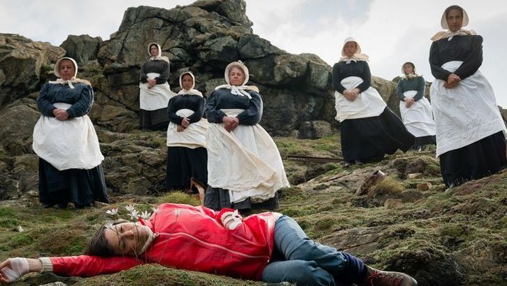 Film still from ENYS MEN: Seven women in black and white robes stand on rocks. In front of them, a woman in a red jumper lies on the ground and looks at a flower directly in front of her.