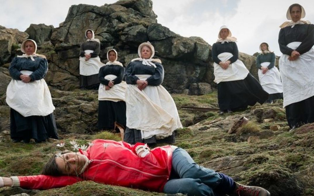 Filmstill aus ENYS MEN: Sieben Frauen in schwarz-weißen Gewändern stehen auf Felsen. Vor ihnen liegt eine Frau in rotem Pullover auf dem Boden und schaut auf eine Blume direkt vor ihr.