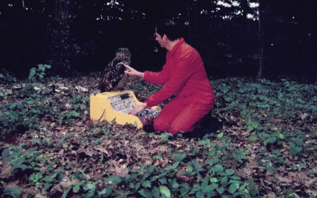 Filmstill aus FELIX IN WONDERLAND: Felix Kubin kniet in einem roten Overall auf dem Waldboden. Vor ihm steht ein Syntheziser, auf dem ein Uhu sitzt.