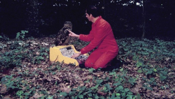 Filmstill aus FELIX IN WONDERLAND: Felix Kubin kniet in einem roten Overall auf dem Waldboden. Vor ihm steht ein Syntheziser, auf dem ein Uhu sitzt.